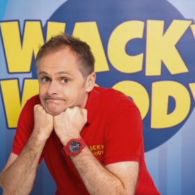 A man wearing a red t-shirt looking puzzled with both hands under his chin. A blue sign in the background with yellow lettering reads Wacky Woody.