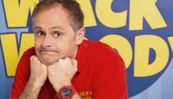 A man wearing a red t-shirt looking puzzled with both hands under his chin. A blue sign in the background with yellow lettering reads Wacky Woody.