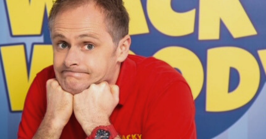 A man wearing a red t-shirt looking puzzled with both hands under his chin. A blue sign in the background with yellow lettering reads Wacky Woody.