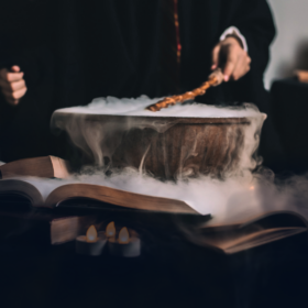 A cauldron bubbling with steam on a table top as somebody stirs it. The cauldron is surrounded by candles.