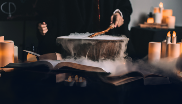 A cauldron bubbling with steam on a table top as somebody stirs it. The cauldron is surrounded by candles.