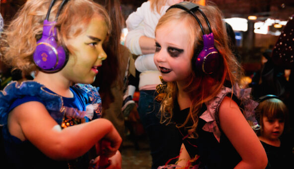 Two young girls dressed in Halloween costume with face paints wearing headphones as if they are enjoying a silent disco.