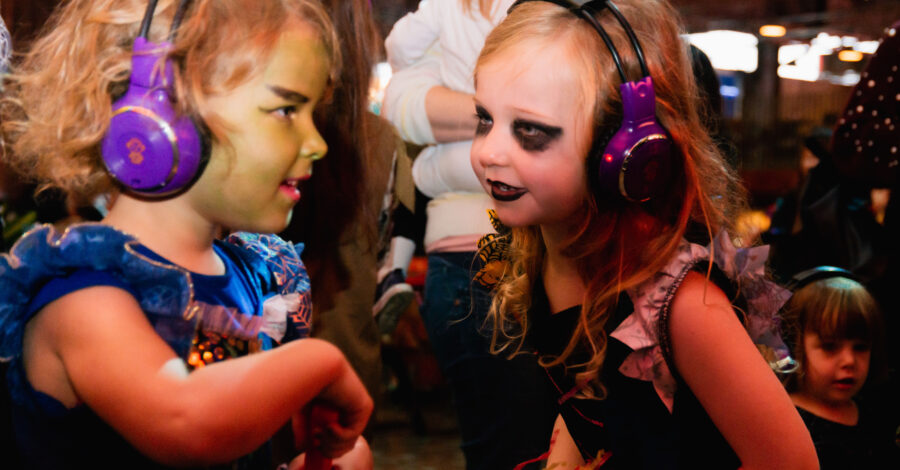 Two young girls dressed in Halloween costume with face paints wearing headphones as if they are enjoying a silent disco.