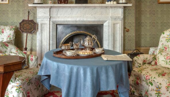 The fireplace and a table with a blue tablecloth on it in the drawing room of Elizabeth Gaskell's House. At the bottom of the image is the text ' 10 years of Elizabeth Gaskell's House'.