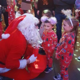 Two small children, dressed in their pyjamas, talk to someone kneeling down who is dressed in a red and white suit and wearing a white beard, like Father Christmas, at the Our Kids Social Xmas Pyjamarama Party.