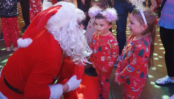 Two small children, dressed in their pyjamas, talk to someone kneeling down who is dressed in a red and white suit and wearing a white beard, like Father Christmas, at the Our Kids Social Xmas Pyjamarama Party.