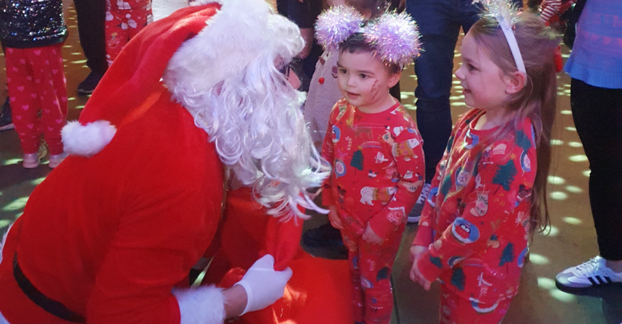 Two small children, dressed in their pyjamas, talk to someone kneeling down who is dressed in a red and white suit and wearing a white beard, like Father Christmas, at the Our Kids Social Xmas Pyjamarama Party.