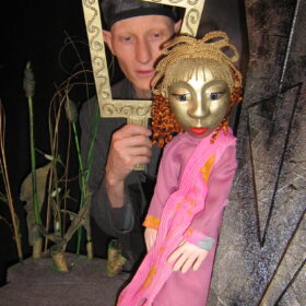 A puppet with a golden face and hair with a performer who stands holding a golden picture frame around his face.