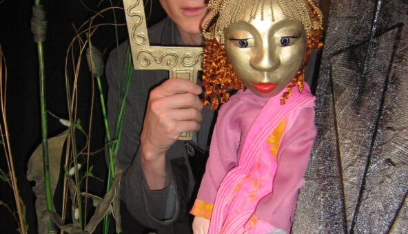 A puppet with a golden face and hair with a performer who stands holding a golden picture frame around his face.