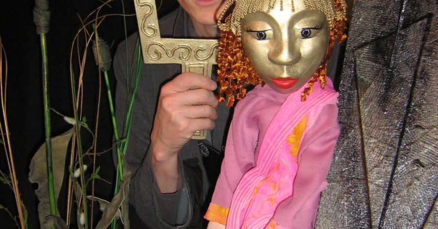 A puppet with a golden face and hair with a performer who stands holding a golden picture frame around his face.