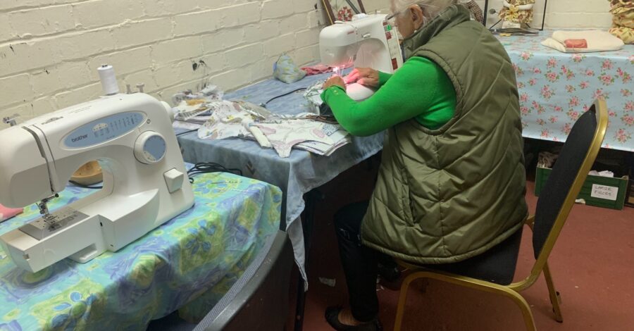 A woman sittng down on a chair at a table using a sewing machine