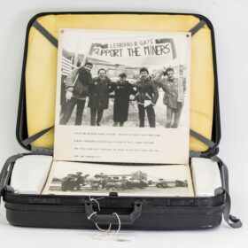 An open black suitcase containing boards from a photographic exhibition, displaying a black and white photograph of five people standing under a banner which reads: ‘Lesbians & Gays Support The Miners’.