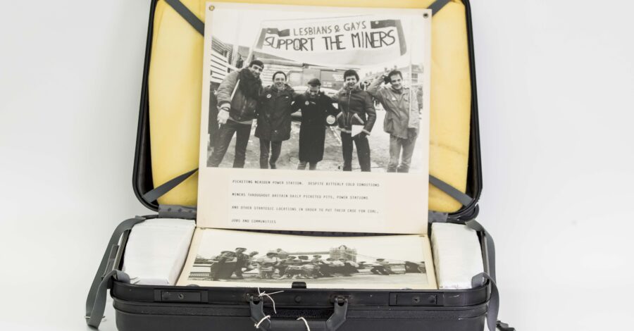 An open black suitcase containing boards from a photographic exhibition, displaying a black and white photograph of five people standing under a banner which reads: ‘Lesbians & Gays Support The Miners’.