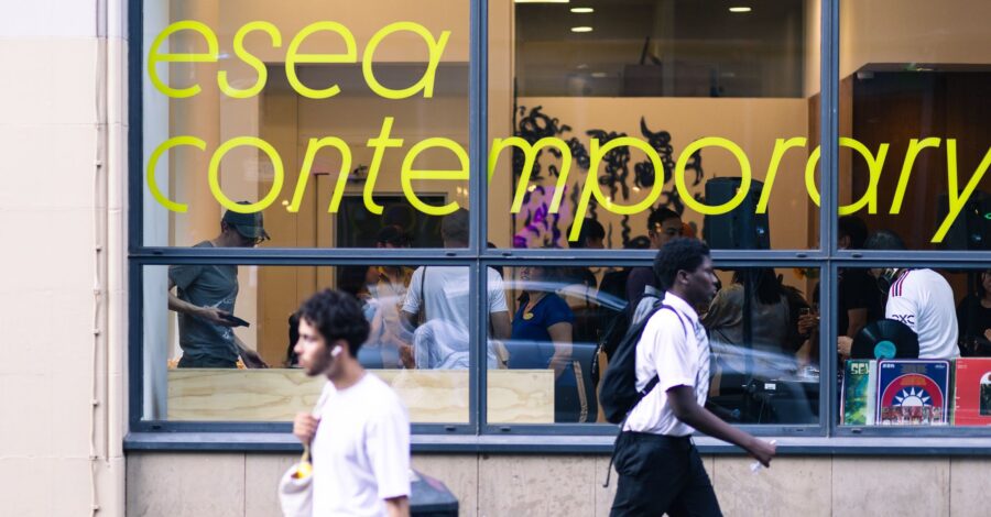 Window front of the gallery with yellow esea contemporary sign, passers by outside, and people inside enjoying the event.