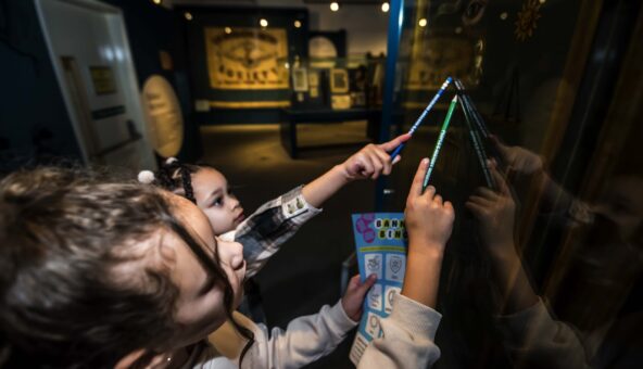 Two young visitors looking up at a banner in a case, holding pencils and an activity sheet.