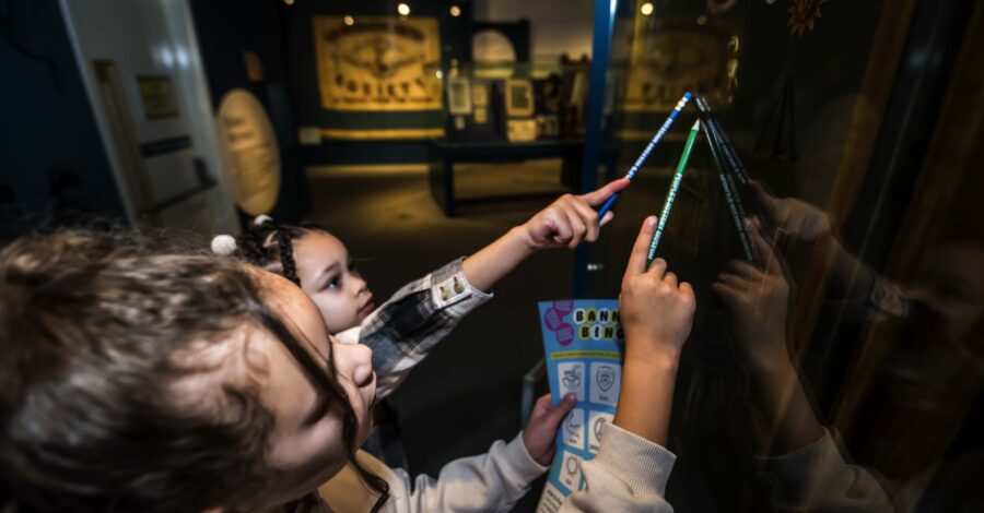 Two young visitors looking up at a banner in a case, holding pencils and an activity sheet.