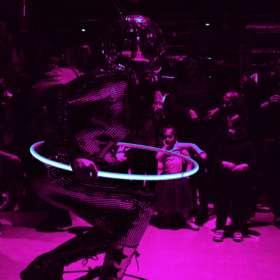 A performer in a reflective bodysuit and disco ball helmet spins a glowing hula hoop under magenta lighting