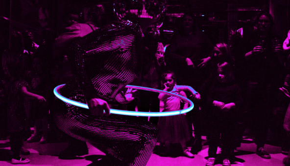 A performer in a reflective bodysuit and disco ball helmet spins a glowing hula hoop under magenta lighting