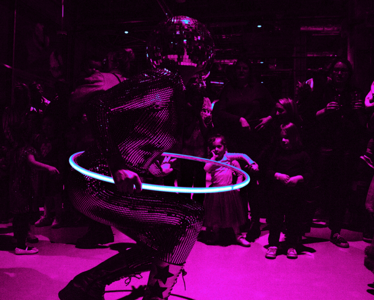 A performer in a reflective bodysuit and disco ball helmet spins a glowing hula hoop under magenta lighting