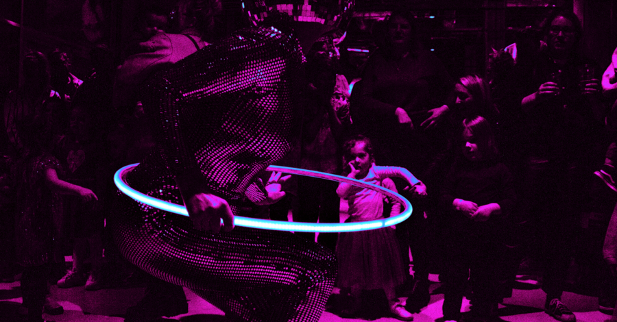 A performer in a reflective bodysuit and disco ball helmet spins a glowing hula hoop under magenta lighting