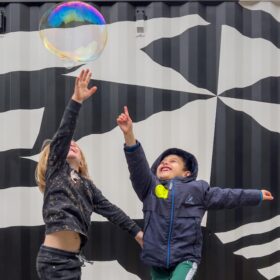 Two small children reaching up to touch a huge bubble.