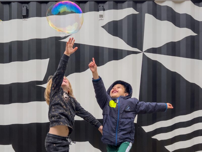 Two small children reaching up to touch a huge bubble.