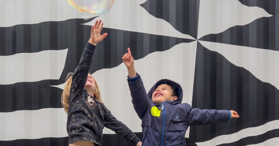 Two small children reaching up to touch a huge bubble.