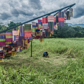 A variety of different coloured cloths hanging from a structure in a grassy field outside. There is a person sat down underneath on the grass, with their knees pulled up, their hands wrapped around their knees, looking down.