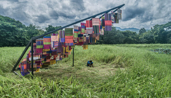 A variety of different coloured cloths hanging from a structure in a grassy field outside. There is a person sat down underneath on the grass, with their knees pulled up, their hands wrapped around their knees, looking down.