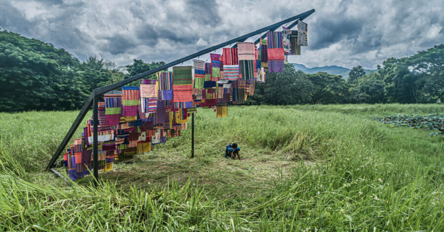 A variety of different coloured cloths hanging from a structure in a grassy field outside. There is a person sat down underneath on the grass, with their knees pulled up, their hands wrapped around their knees, looking down.