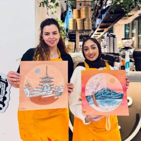 Two people, who are smiling and wearing orange aprons, hold up their Japanese-inspired artwork.