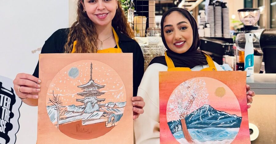 Two people, who are smiling and wearing orange aprons, hold up their Japanese-inspired artwork.