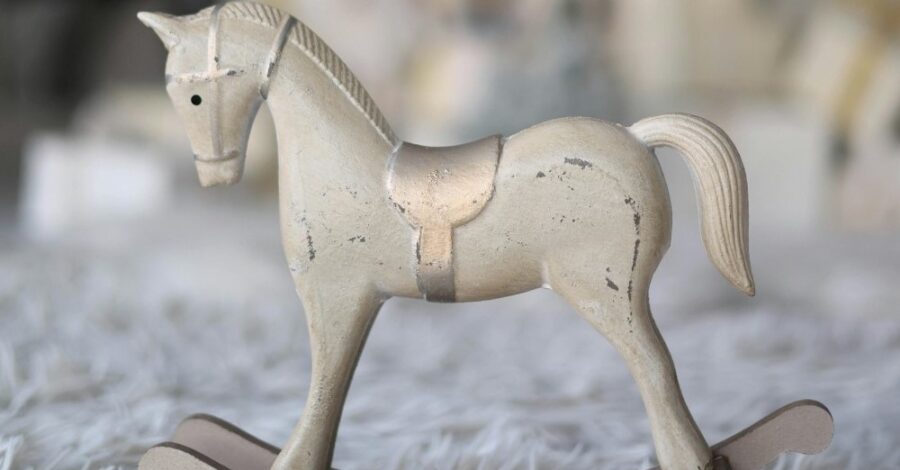 A small white wooden rocking horse with a gold saddle. The toy is placed on a fluffy grey carpet.