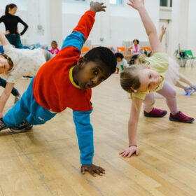 Two young children pose with two feet and one hand on the floor while the other arm is extended towards the ceiling in a studio.