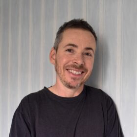 A photograph of Jay Prosser. A middle-aged man with short dark hair and brown eyes. He is smiling. He's got a short-trimmed beard and is wearing a black T-shirt.