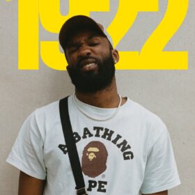 A man wearing a t-shirt and a cap stands infront of the numbers 1922, which are on the wall behind him in yellow.