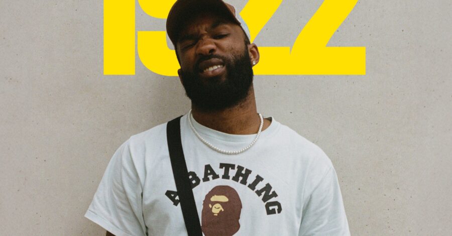 A man wearing a t-shirt and a cap stands infront of the numbers 1922, which are on the wall behind him in yellow.