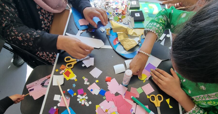 Birds eye view of a table filled with coloured paper, pens and scissors where children are crafting.