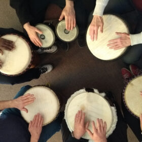 Image from above of 6 pairs of hands playing small drums