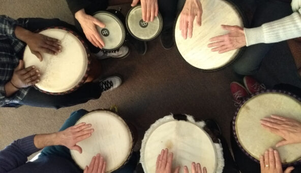 Image from above of 6 pairs of hands playing small drums