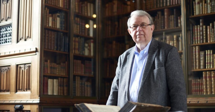 Photograph of Professor Philip Alexander. It shows an older man with grey hair and glasses standing in front of an open book in a historic library with wooden bookshelves in the background.