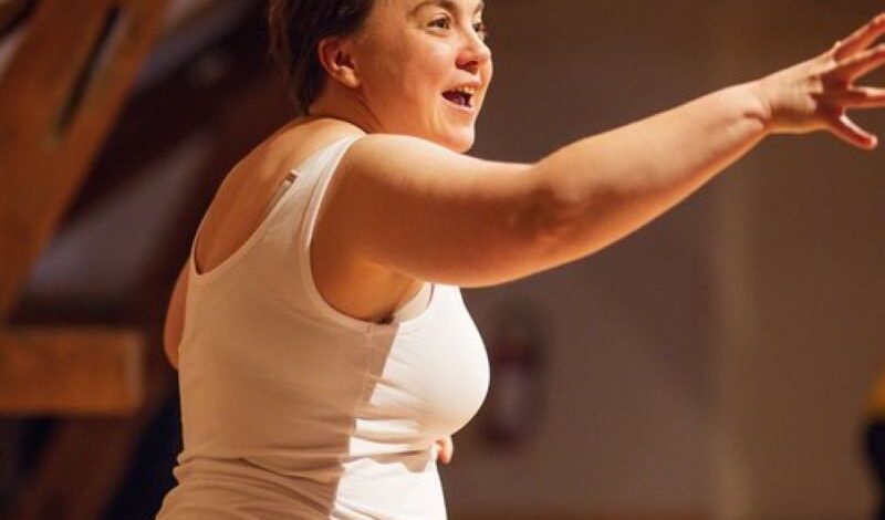 A woman stands performing, arm outstretched