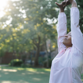 A woman in a green space stretching upwards as the sun shines between a tree.