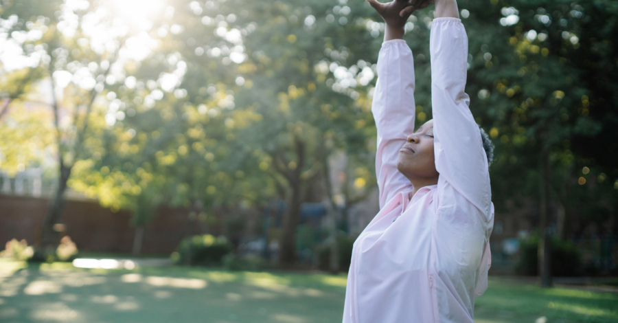 A woman in a green space stretching upwards as the sun shines between a tree.