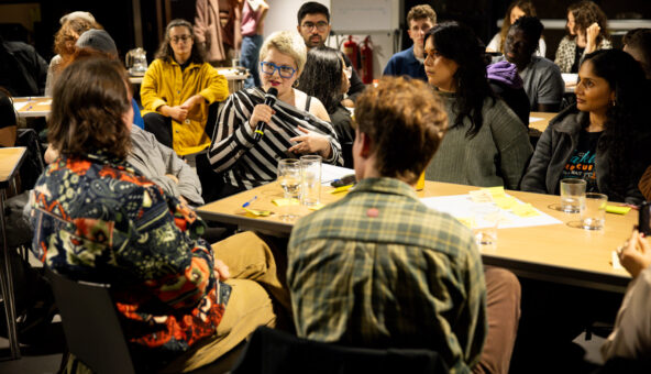 Someone speaking into a microphone sat at a table surrounded by others who are listening.