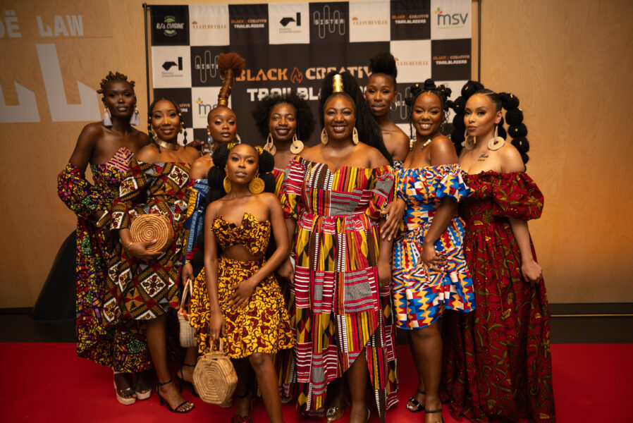 A group of women with brightly coloured dresses stood together, smiling, as if a photograph is being taken.