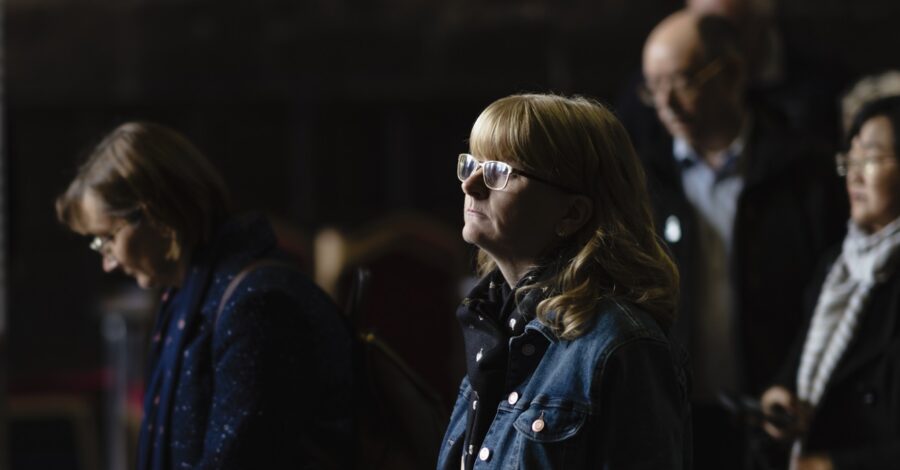 A lady with glasses, light brown hair, wearing a blue jacket stood staring at something forward in a dark room.