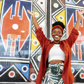 A lady stands infront of a wall painted with vibrant patterns, with her arms raised up and she is smiling.