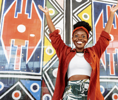 Woman in front of a wall painted with vibrant patterns, with her arms raised and she is smiling.