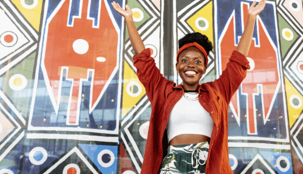 A lady stands infront of a wall painted with vibrant patterns, with her arms raised up and she is smiling.